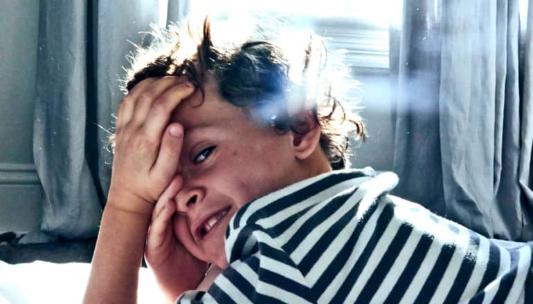 A young boy smiles and touches his face while laying on a bed