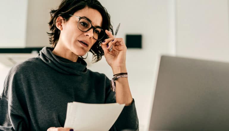 serious person holds papers and glasses in front of laptop