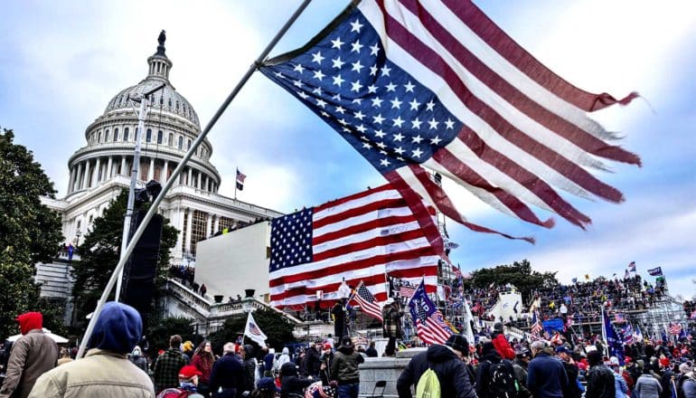 Rioters storm the US capitol building holding American flags, with a tattered flag waving in the foreground