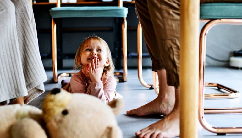 A little girl smiles while laying under a table as her parents work from home