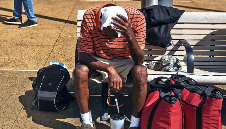 A man sits on a bench surrounded by baggage while he covers his face with a cloth