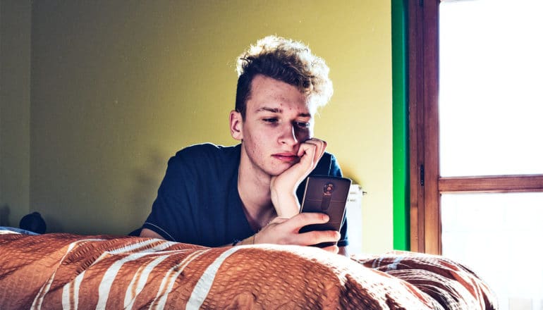 A young man looks at his phone while laying on his bed with his chin resting on his hand