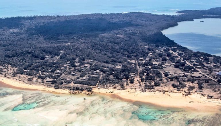 An aerial view of a Tongan island covered in dark ash, including homes near the beach