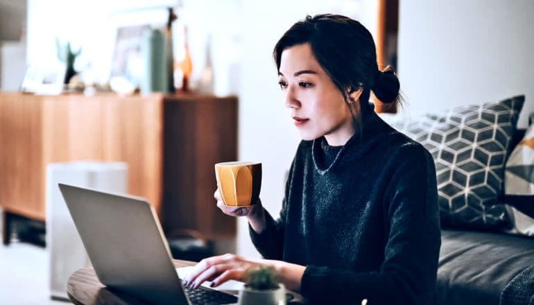 A woman works from home on a laptop while holding a cup of coffee