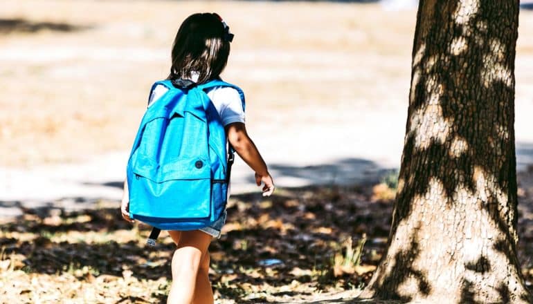 A young girl walks to school through a field while carrying a blue backpack