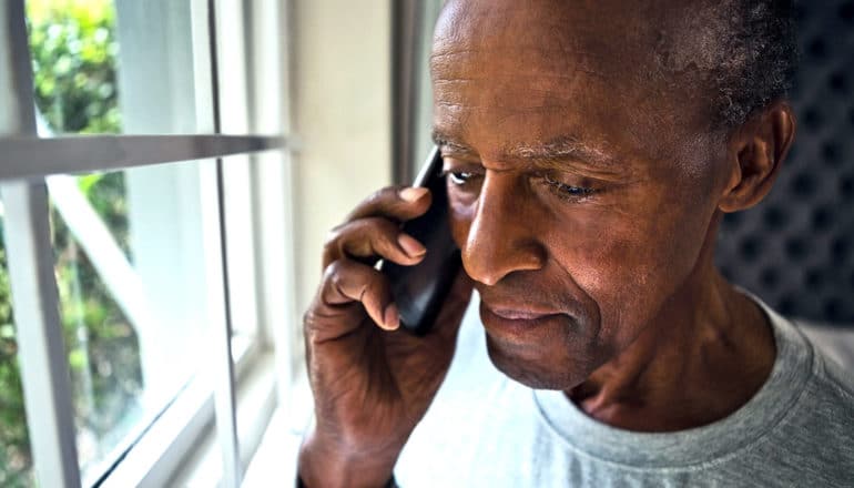 An older man speaks on the phone while standing near a window in his home