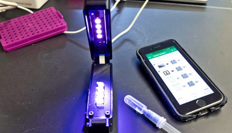 A smartphone sits next to the black detector, which has several purple lights shining on it