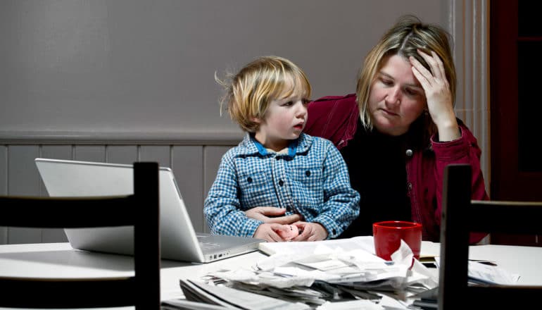 upset adult holds child at table with laptop, paperwork, and mug