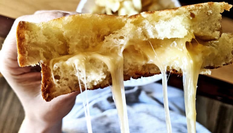 A person holds a broken grilled cheese sandwich with cheese stretching off camera