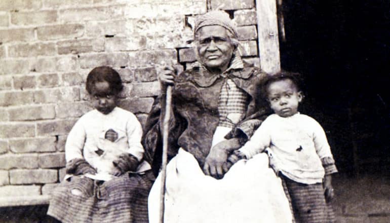 small, serious children on either side of elderly woman holding cane