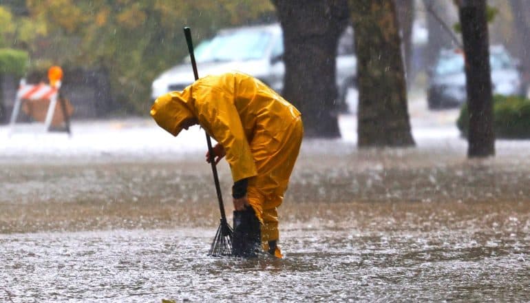 A worker in yellow rain gear bends over to clear a drain in a flooded street