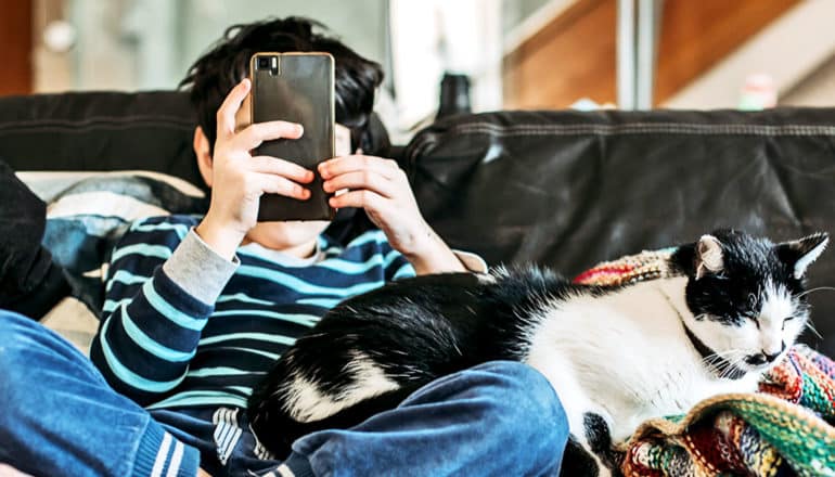 A young boy sits on a couch next to a cat while looking at a phone