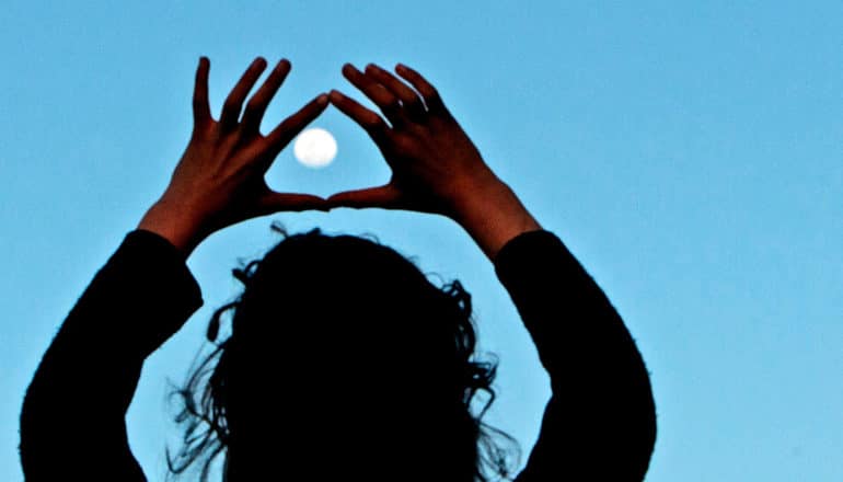 A woman holds her hands up to frame the moon in the evening sky