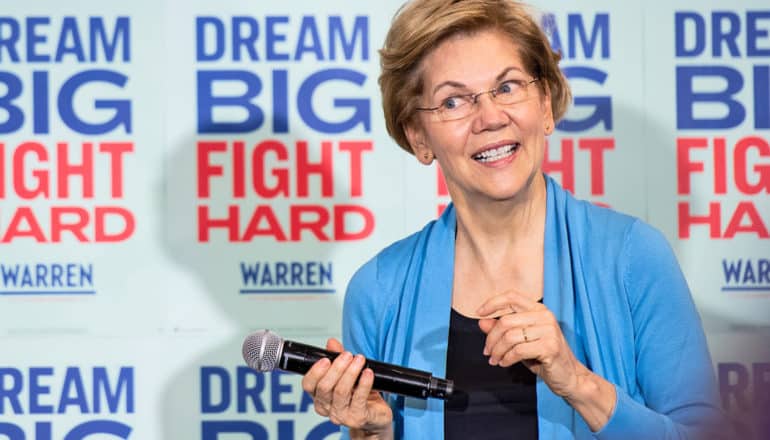 exasperated Elizabeth Warren holds mic in front of "dream big fight hard" signs