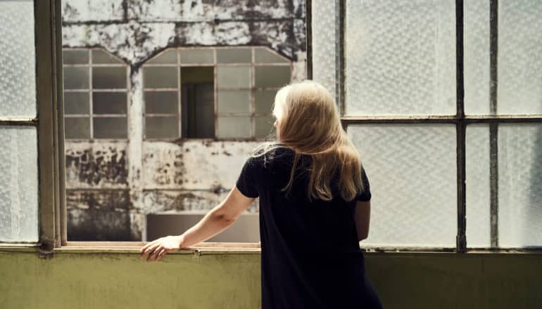 person with long hair looks out of dingy window