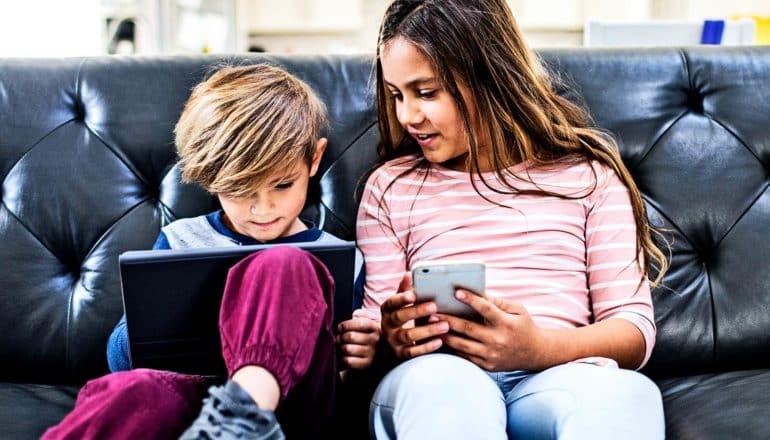 A young boy and older girl sit on a couch while using a tablet and smartphone