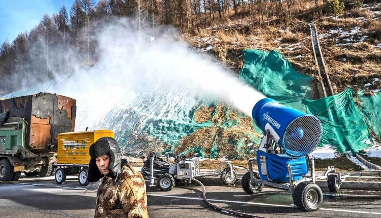 A worker stands next to a snow machine spraying artificial snow onto the side of a hill