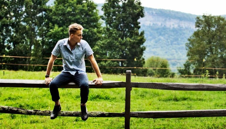 A young man sits on a fence outside with trees in the background