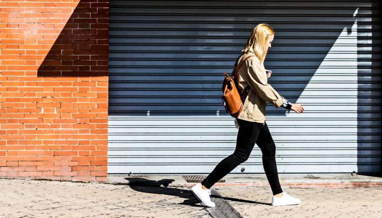 A young woman walks on a sidewalk past a closed metal door in a brick building