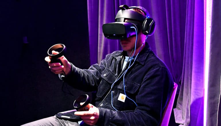 A man uses a virtual reality headset and controllers against a curtain cast in purple light