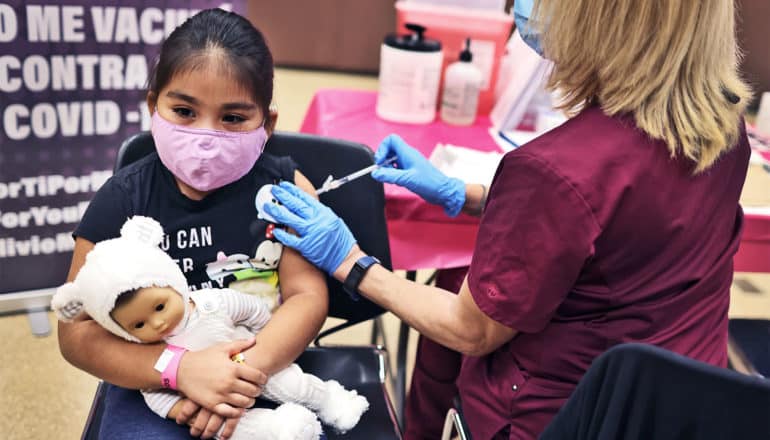 A young girl gets a covid-19 vaccine while holding a doll in a onesie