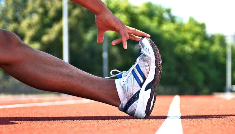 A man reaches towards the toe of his running shoe while stretching on a red track