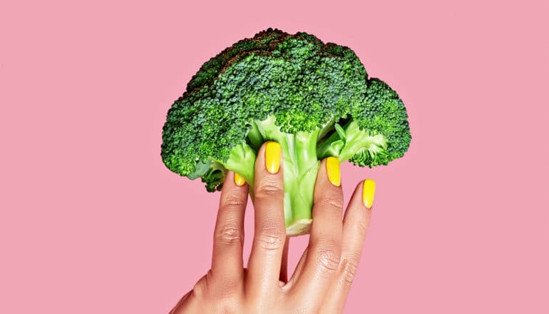 A woman with painted yellow nails holds a piece of broccoli against a pink background