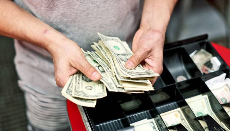 hands hold stack of $1 bills over cash register drawer