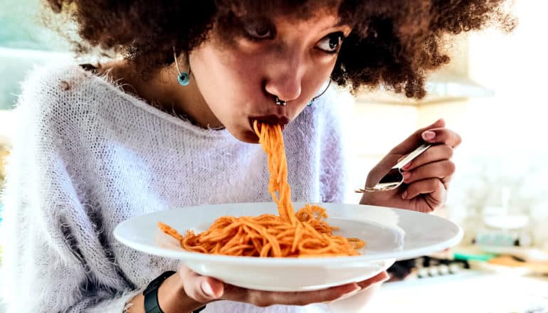 A woman eats a big mouthful of spaghetti while sitting in her kitchen