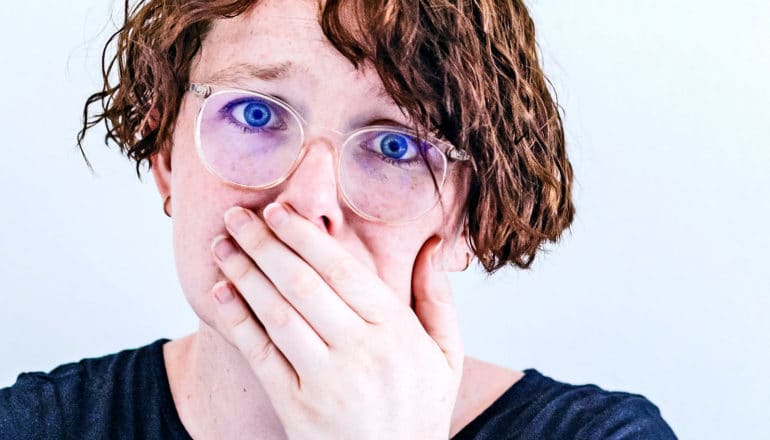 A young woman with glasses looks scared as she covers her mouth with her hand