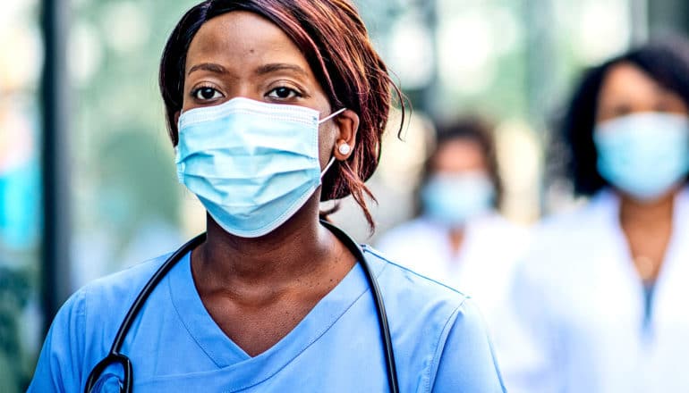 A nurse wearing scrubs and a medical mask walks outside a hospital