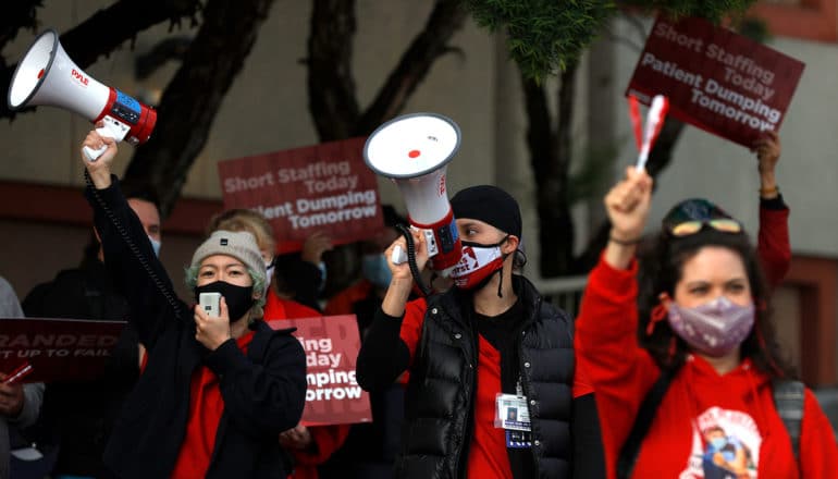 three people in masks, two will bullhorns, wear red and black. Signs behind them read " short staffing today patient dumping tomorrow"