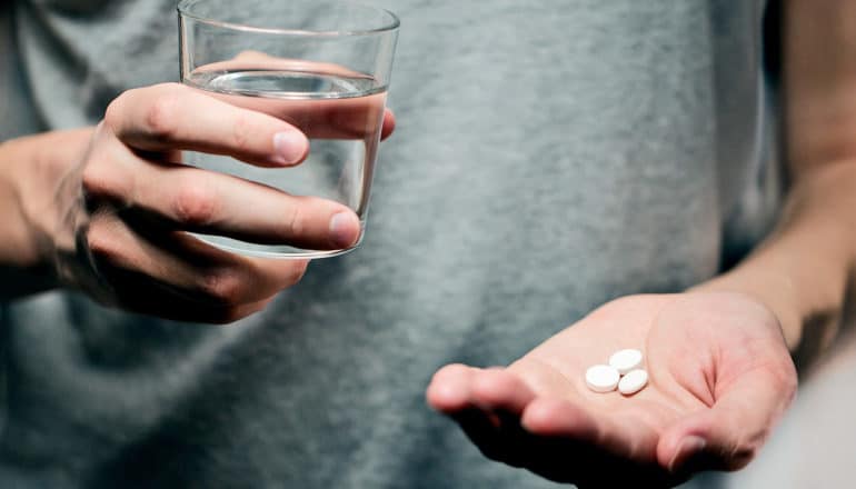 A man holds 3 pills in one hand and a glass of water in the other