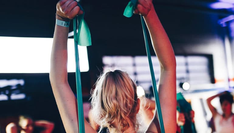 A woman pulls a green resistance band above her head while in an exercise class