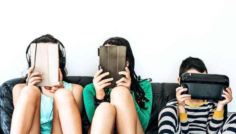 Three young teens, two girls and a boy, sit on a couch with tablet computers and gaming consoles covering their faces