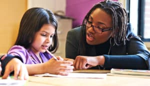 A teacher helps a student working at a classroom desk