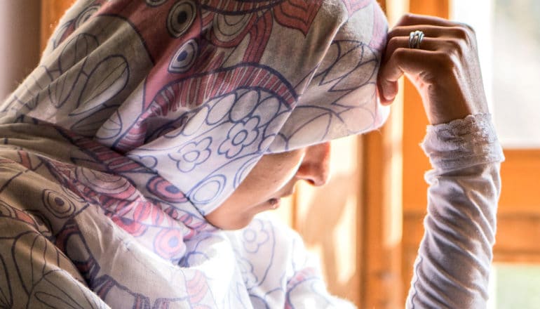 A woman sitting by a window wearing a headscarf rests her head on her hand and looks down