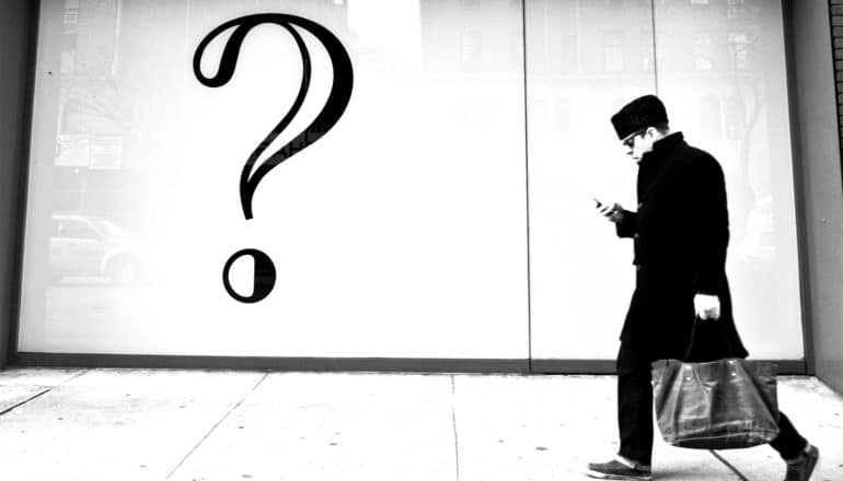 A man walks down the sidewalk looking at his phone while passing a store with a large question mark hanging in the window