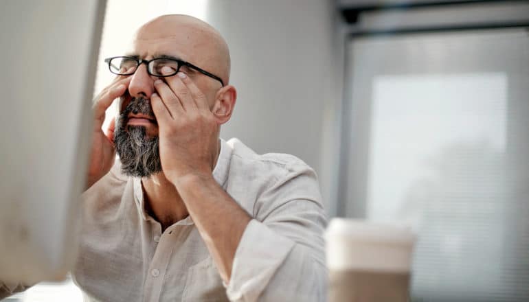 adult with gray beard, glasses, bald head, rubs eyes in front of computer