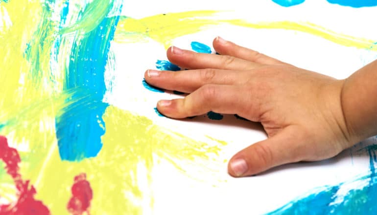 A young child finger painting places a handprint in blue paint on a white canvas covered in other colors of paint