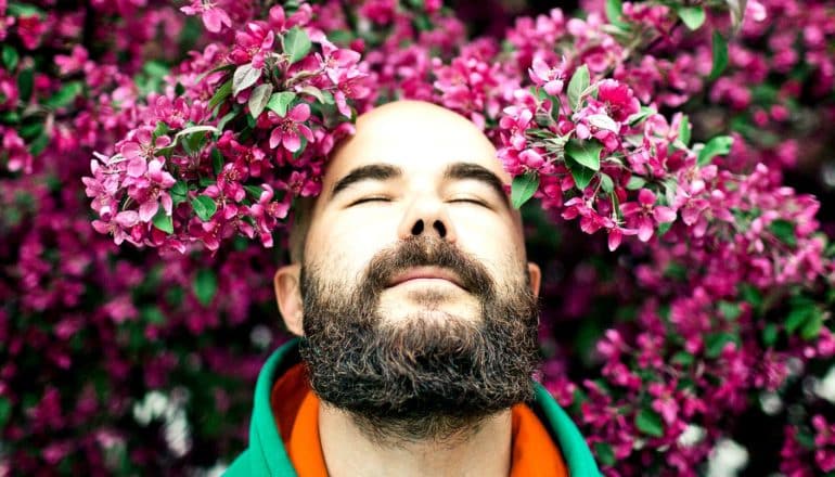 A bald man breathes deeply with his eyes closed as he's surrounded by purple flowers on trees