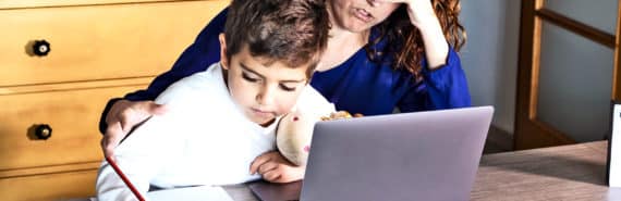 A mother puts her hand over her face as she sits with her son, working on a laptop for school.