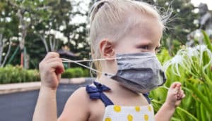 A little girl holds a gray medical mask over her face while standing in a park.