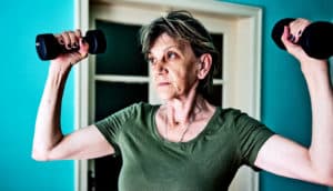 A woman lifts weights above her head in a blue room with a window behind her.