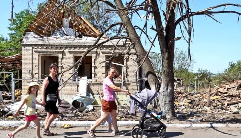 A family with a young child and baby in a stroller walk past a bombed out house and rubble.