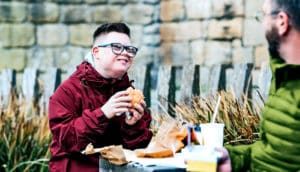 A boy with Down syndrome eats a burger while smiling and sitting with his dad.