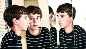 A teen boy looks in a mirror, with several other reflections of himself appearing in nearby mirrors.