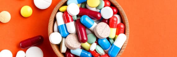 A bunch of different pills in a small bowl, spilling over onto an orange background.