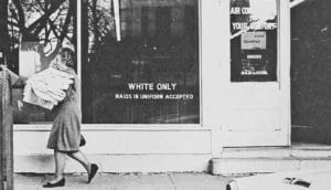 black and white photo of person walks by "white only maids in uniform accepted" written on window