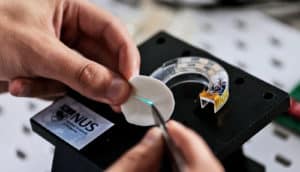 A researcher touches a small phosphor-infused contact pad with a tool, which prompts blue light to show on the pad. The mouthguard device, which is clear plastic and contains electronic components, sits below on a black surface.